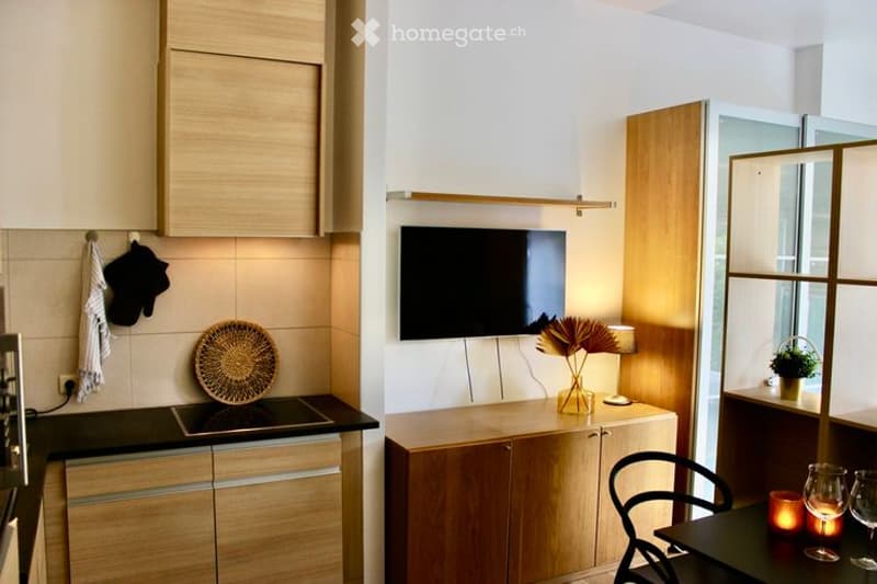 Modern kitchen and living area with light wood cabinets, induction stove, wall-mounted TV, small dining table with chairs, decorative vase, candle, and wine glasses.