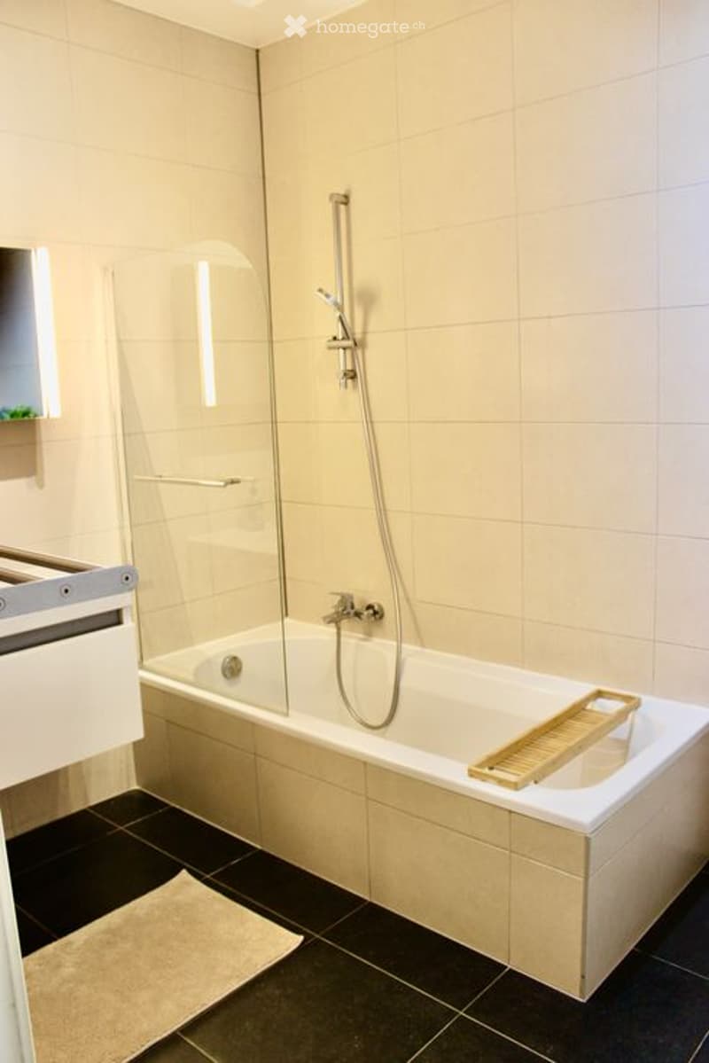 Modern bathroom with a large bathtub, glass shower enclosure, and a wooden tray inside the tub. The walls are tiled in light beige, and the floor is black.