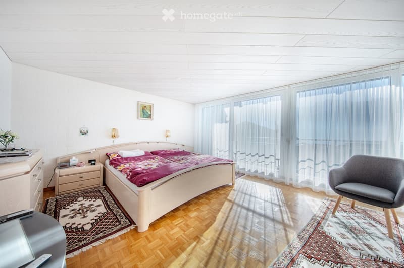 Bright bedroom with wooden flooring, a double bed with a floral duvet, a nightstand with a lamp, a grey armchair, and large windows with sheer curtains.