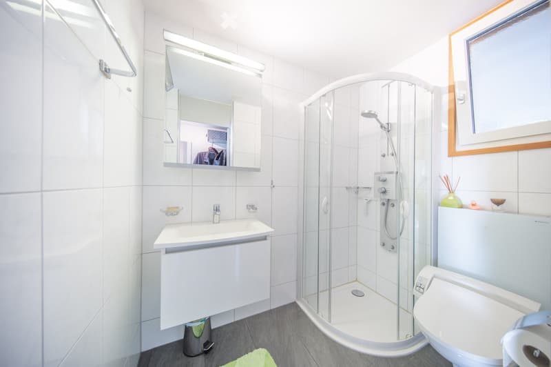 Modern bathroom with white tiles, a glass-enclosed shower, a sink with a mirror, and a toilet. Includes a small cabinet and a towel on the floor.
