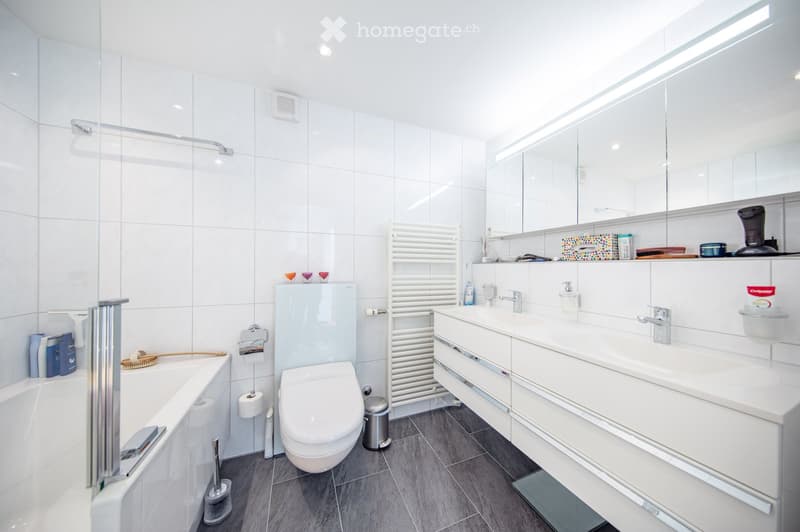 Modern bathroom with white tiles, a bathtub, toilet, double sink, large mirror, and a towel rack. The space is bright and clean with a glass-enclosed shower area.