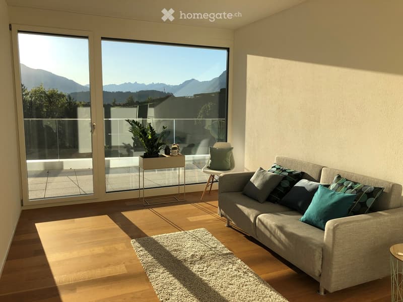 Modern living room with large windows offering a scenic mountain view, featuring a grey sofa with multiple throw pillows, a small coffee table, a white chair, a shaggy rug, and a potted plant on a side table.