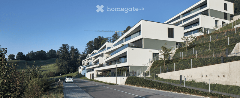 Modern multi-story residential building with white and grey facade, large windows, and balconies, situated on a hillside with landscaped surroundings.
