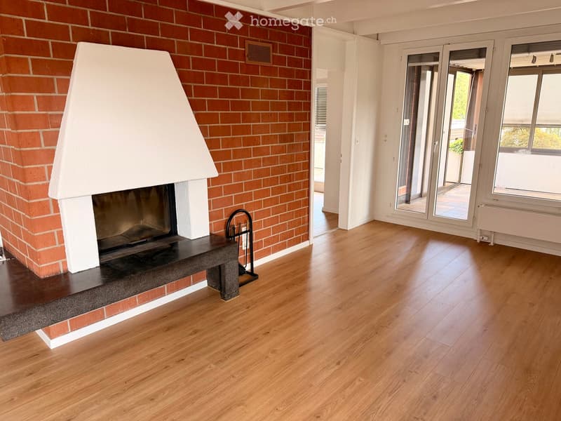 Empty room with wooden flooring, a brick fireplace with a white mantel, and large windows with sliding glass doors leading to an outdoor area.