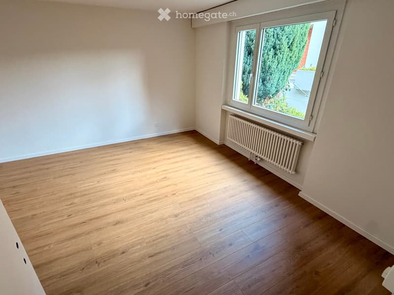 Empty room with wooden flooring, white walls, and a large window with a radiator below it.
