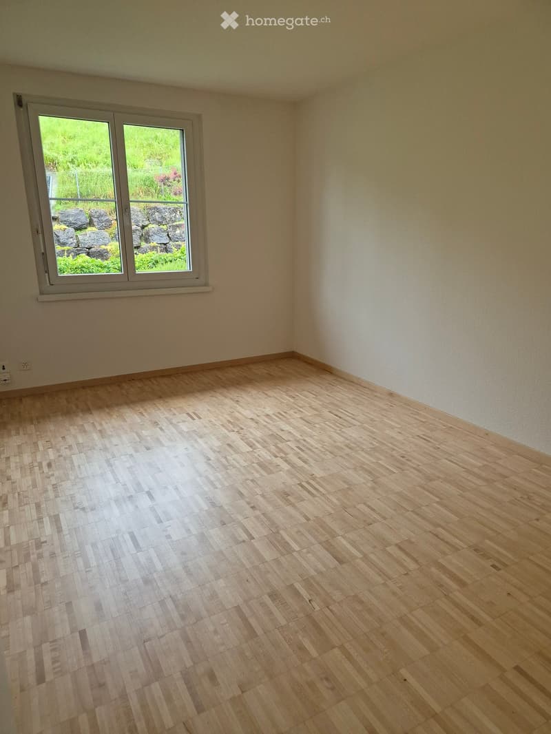 Empty room with light wooden flooring and white walls, featuring a large window with a view of greenery and rocks outside.