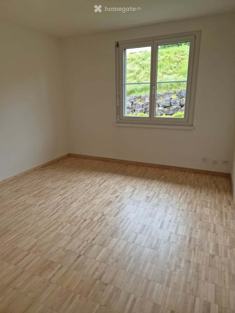 Empty room with wooden flooring and a large window showing a view of greenery and rocks outside.