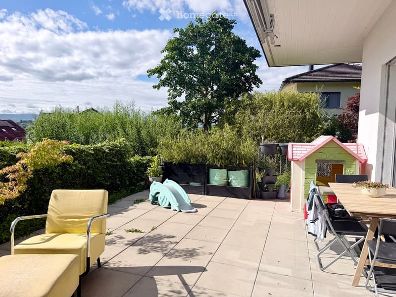 A sunny patio with a yellow chair, a small wooden table with chairs, a colorful toy house, and several potted plants. The area is surrounded by greenery and has a view of the landscape.