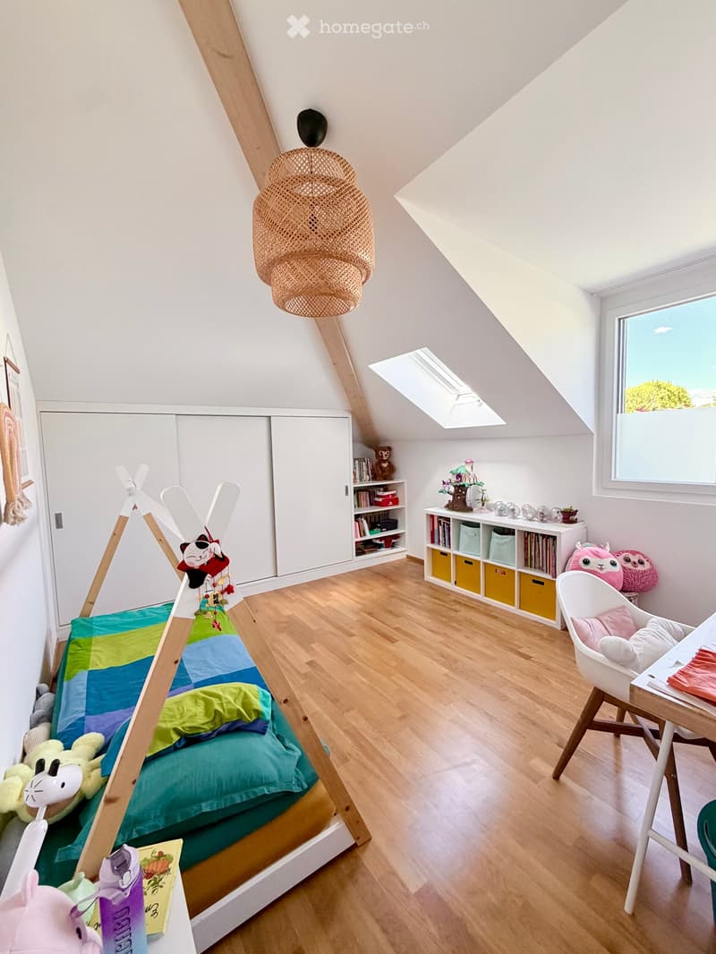 Bright, modern attic bedroom with wooden floors, slanted ceilings, and large windows. Features a colorful bed, a white chair, a small table, a bookshelf with storage boxes, and decorative toys.