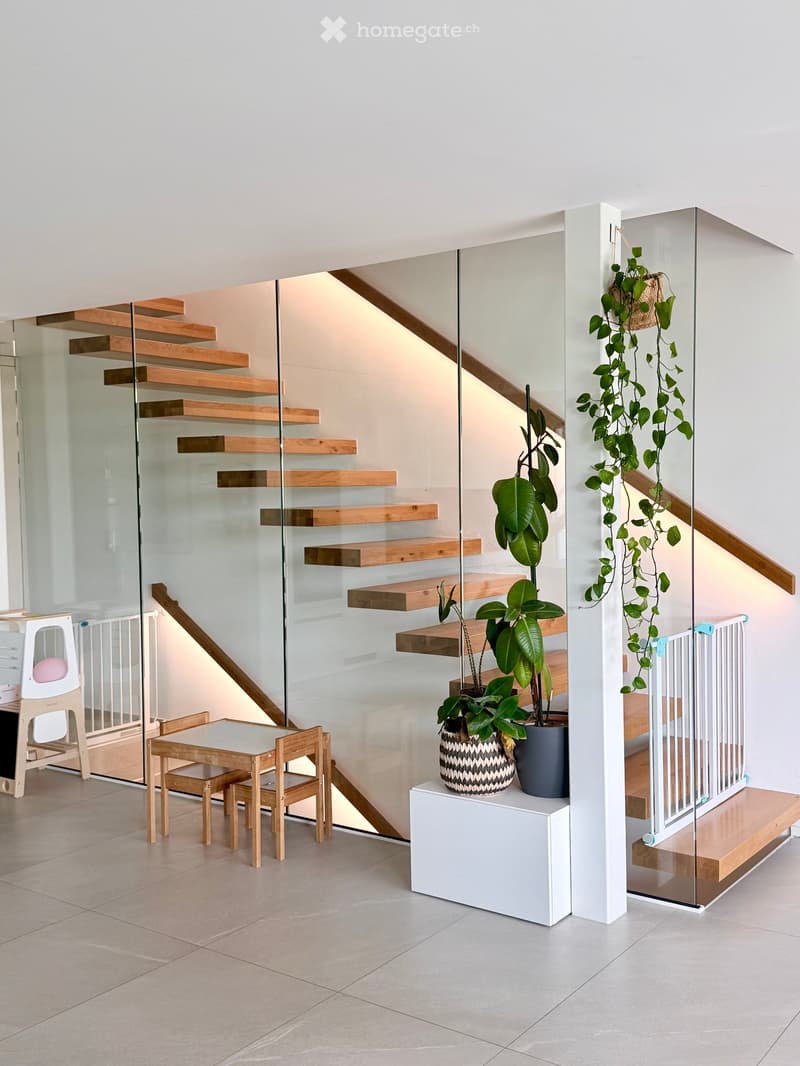 Modern interior with a glass staircase, small wooden table and chairs, baby chair, and potted plants on a white stand.