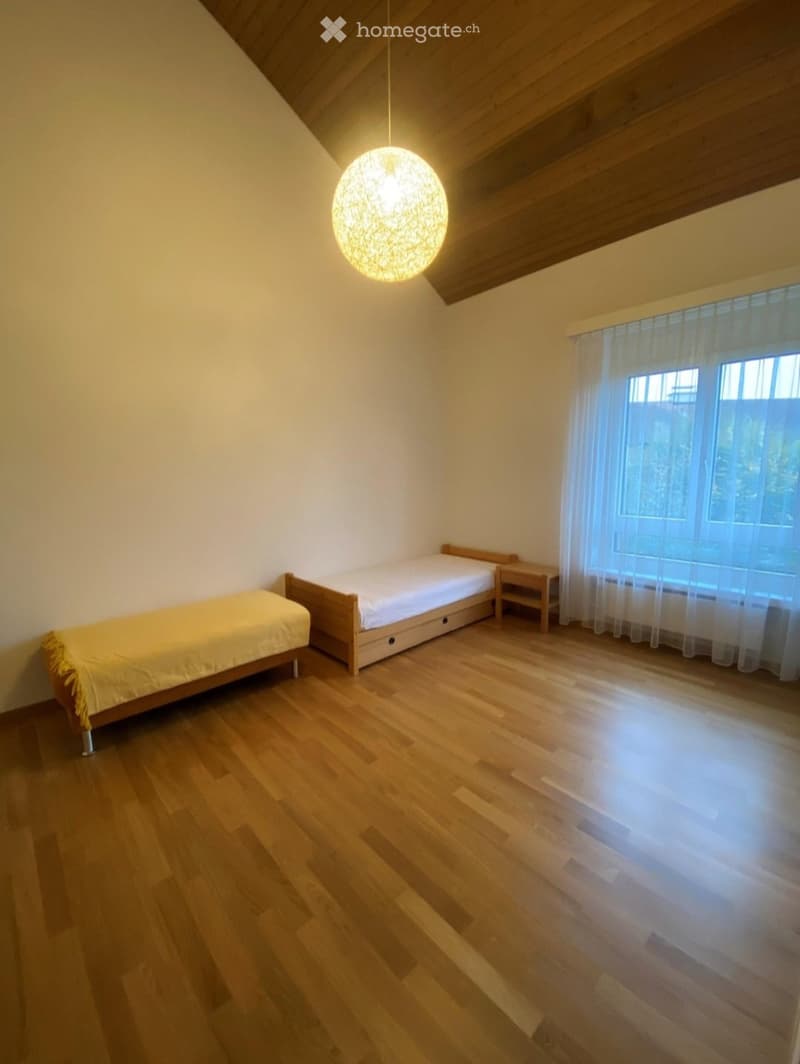 Cozy bedroom with wooden floors, two single beds, a nightstand, and a large window with sheer curtains.