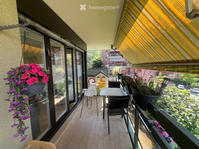 Balcony with tiled floor, glass sliding doors, outdoor furniture including a table and chairs, potted plants, and hanging flower basket.