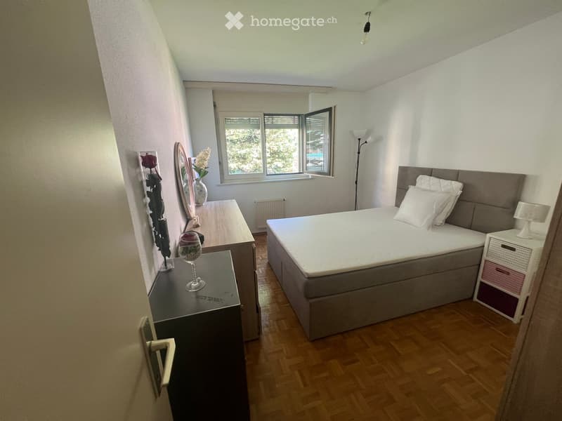 Bedroom with a double bed, nightstand, mirror, vase with flowers, and a drawer unit. The room features wooden flooring and large windows with blinds.