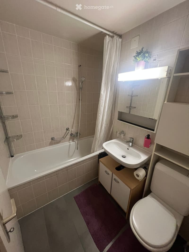 Bathroom with a bathtub, sink, toilet, mirror, and cabinet. Features a purple bath mat and a window with a curtain.