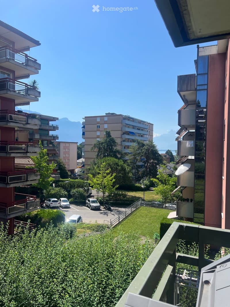 Balcony view overlooking a residential area with multiple apartment buildings, parked cars, and a clear view of mountains in the distance.