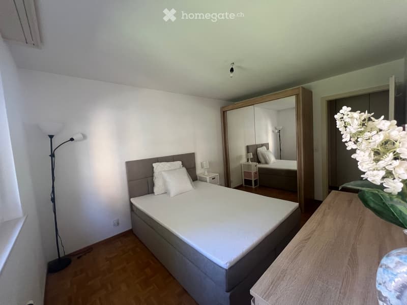 Modern bedroom with a large bed, nightstand, mirror, and a vase of white flowers on a wooden table.