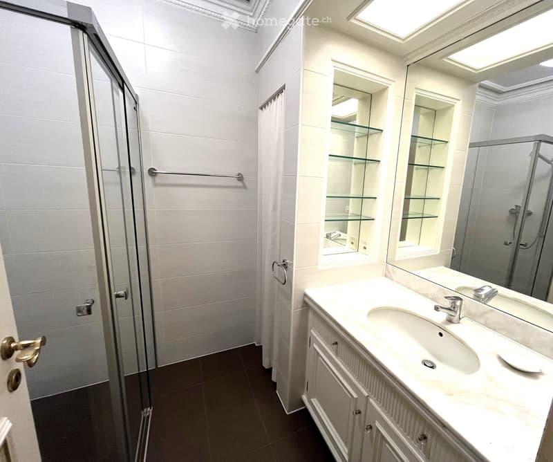 Modern bathroom with white tiles, a glass-enclosed shower, a white vanity with a sink and mirror, and glass shelves.