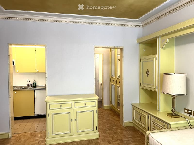 A furnished room with light yellow cabinets, a sink, a small table, and a lamp on a wooden floor.