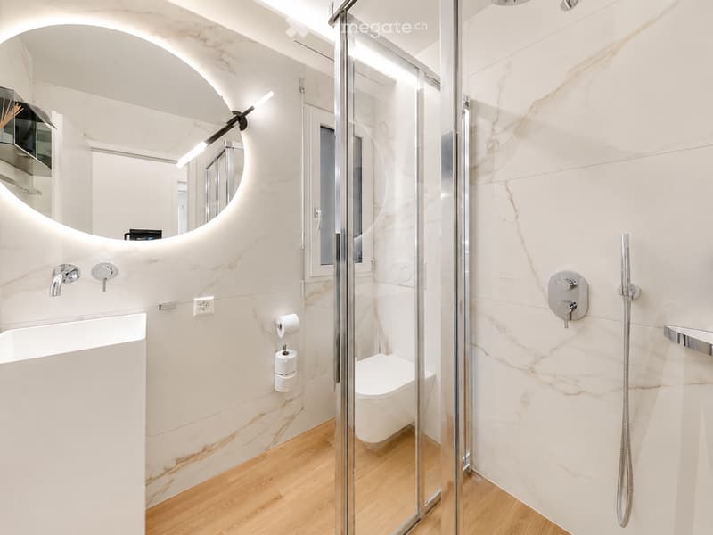 Modern bathroom with marble walls, a glass-enclosed shower, a toilet, a sink with a modern faucet, and a round mirror above the sink.