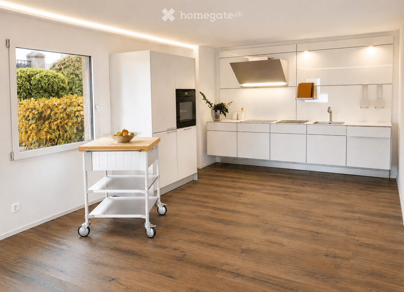 Modern kitchen with white cabinets, stainless steel appliances, wooden floor, and a cart with a bowl of fruit. Large window offers a view of greenery outside.