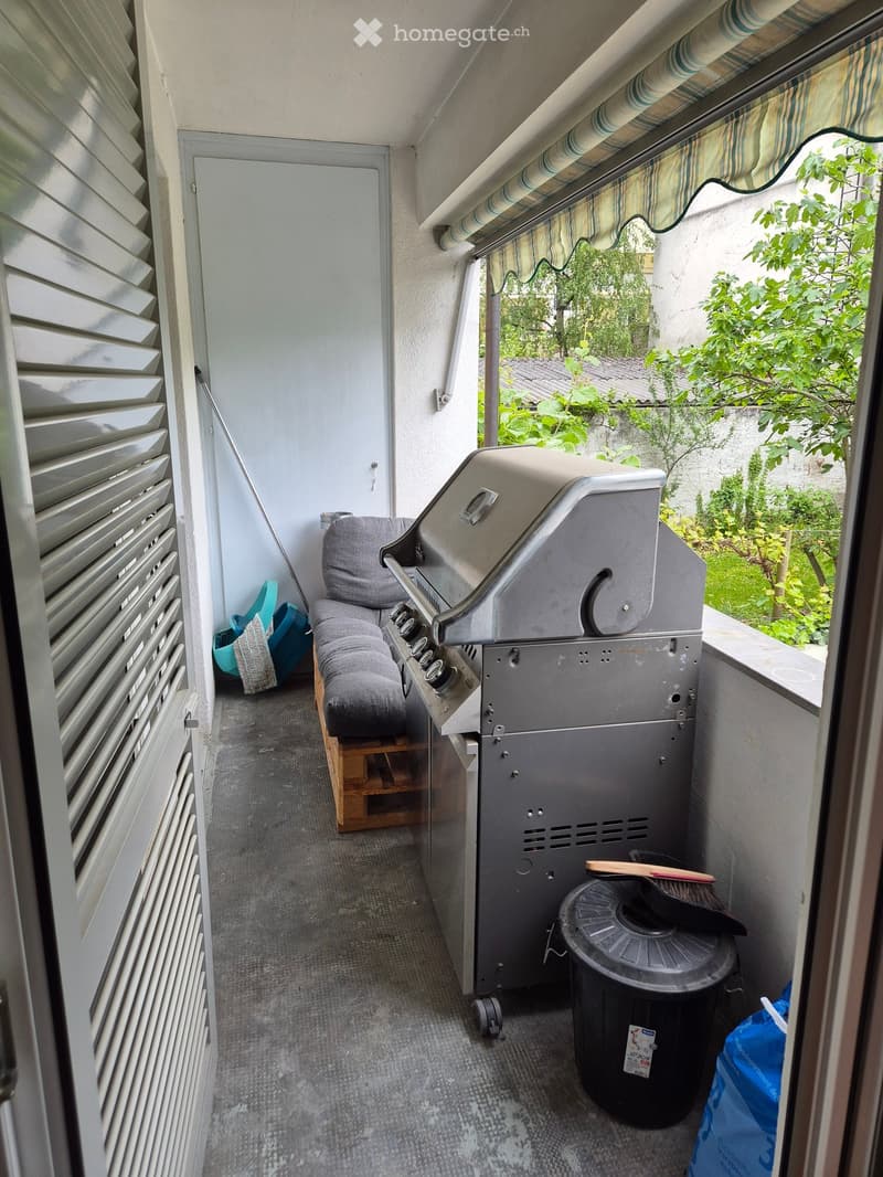 Balcony with a grill, a small bench, and a trash can, featuring a striped awning and a view of greenery.