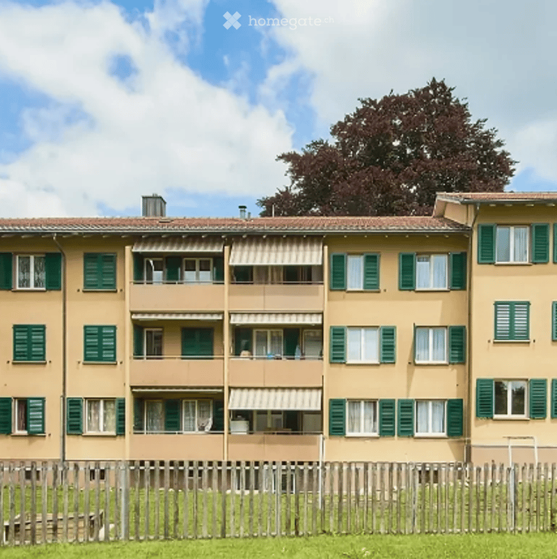 Multi-story residential building with beige facade, green shutters, and multiple windows. Balconies with awnings are visible on each floor.