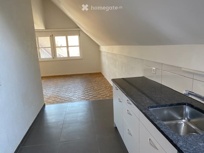 Empty kitchen with white cabinets, black countertop, stainless steel sink, and tiled flooring. The room has a slanted ceiling and a window with multiple panes.