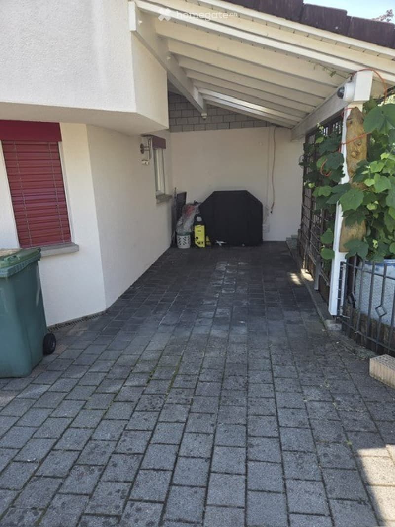 A paved outdoor area with a covered walkway, green trash bin, and a barbecue grill.