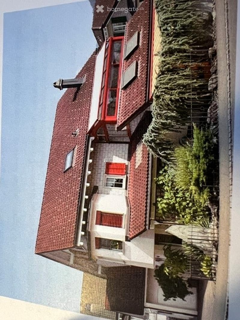A multi-story residential building with a red-tiled roof, white walls, and multiple windows. The structure includes a balcony area with greenery and a small garden space.