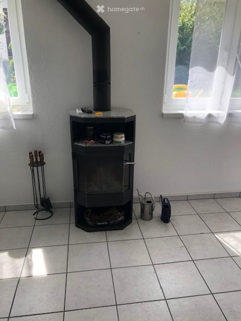 A tiled room with a modern black wood-burning stove, a firewood stand, and a can near the stove.