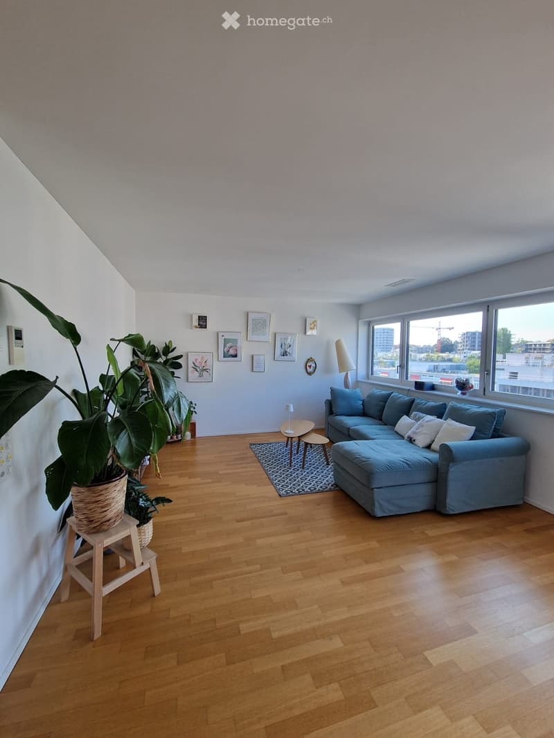 Modern living room with light wooden flooring, large windows, a blue sectional sofa, a small coffee table, a plant in a woven basket, and several picture frames on the wall.