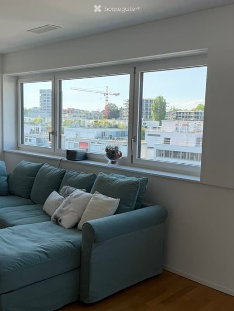 Modern living room with large windows offering a city view, a blue sectional sofa with white throw pillows, and a small flower pot on the windowsill.