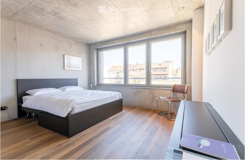 Modern bedroom with a large bed, wooden flooring, large windows, a simple chair, and a small table with a brochure on it.