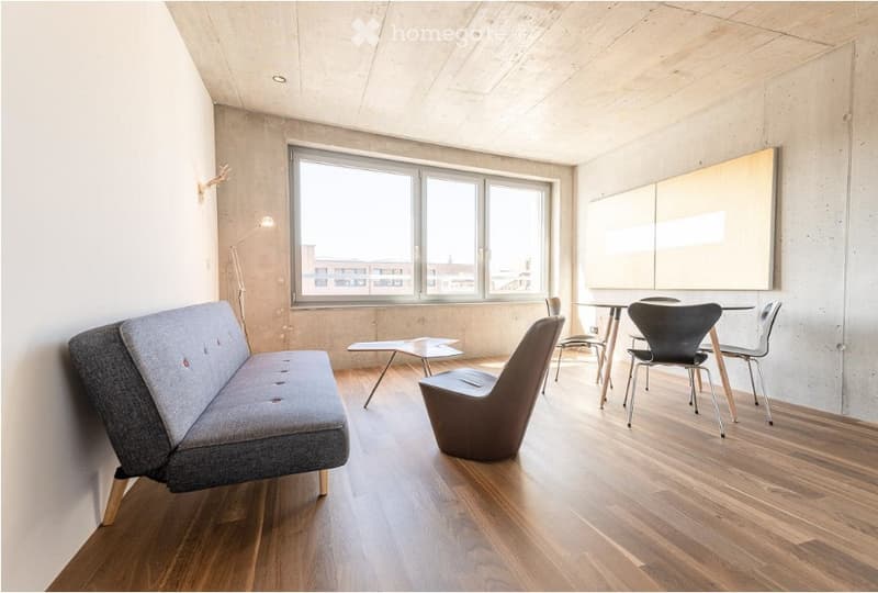 Modern, minimalist living space with wooden flooring, large windows, grey sofa, sleek chair, small table, and modern lamp.
