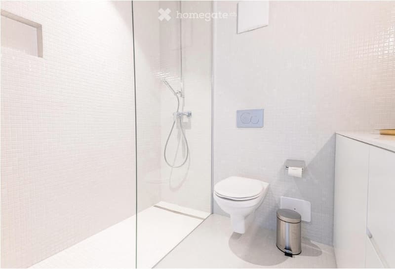 Modern, compact bathroom with white tiled walls and floor, featuring a toilet, a glass-enclosed shower, and a small trash can.