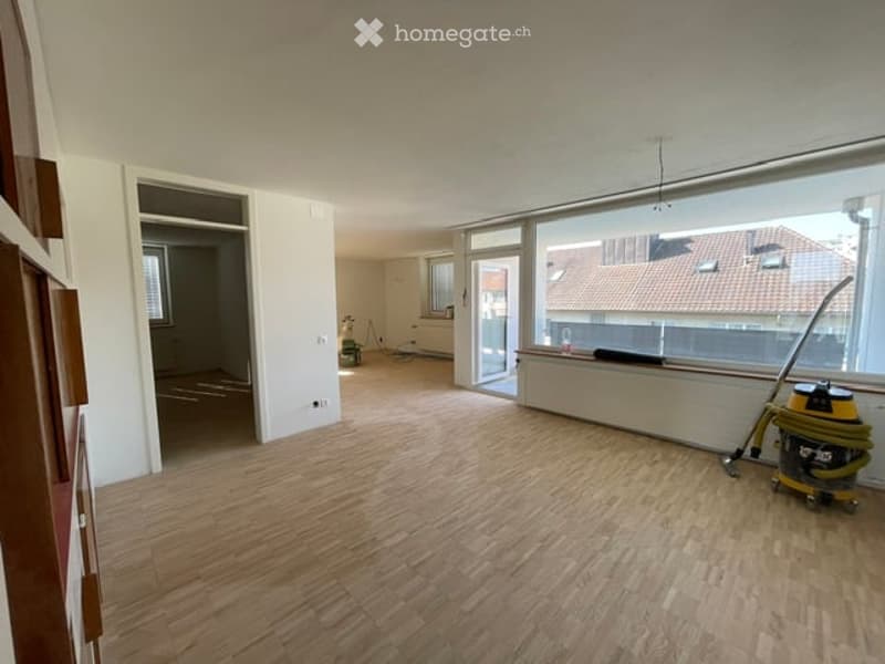 Empty room with wooden flooring, large windows, and a vacuum cleaner in the corner.
