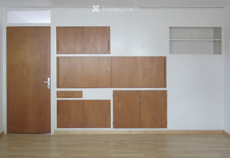 Empty room with wooden floor, white walls, and built-in wooden cabinets with drawers and shelves. A wooden door is visible on the left side.
