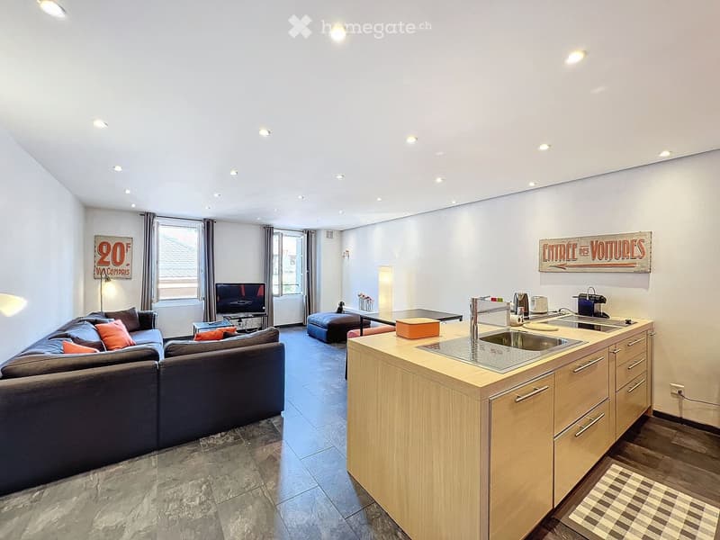 Open-plan living and kitchen area with a sectional sofa, coffee table, kitchen island with sink and appliances, and decorative artwork on the walls.