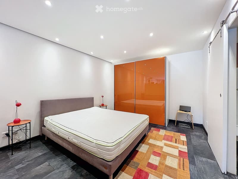 Modern bedroom with a large bed, orange wardrobe, small table with a lamp, and a small desk with a screen.