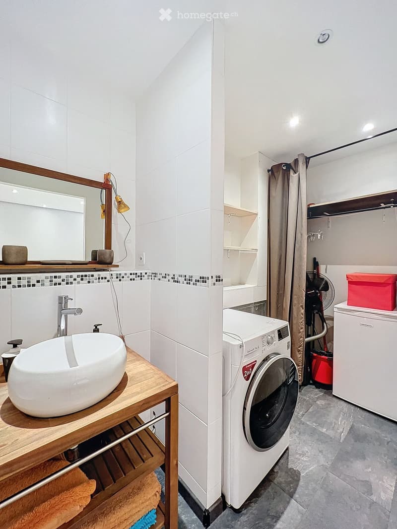 Modern bathroom with a white sink, wooden shelf, and a separate laundry area featuring a washing machine and storage boxes.