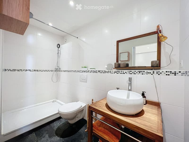 Modern bathroom with white tiles, a bathtub, toilet, sink, and a large mirror with wooden frame. Includes a wooden vanity with a sink and a towel underneath.