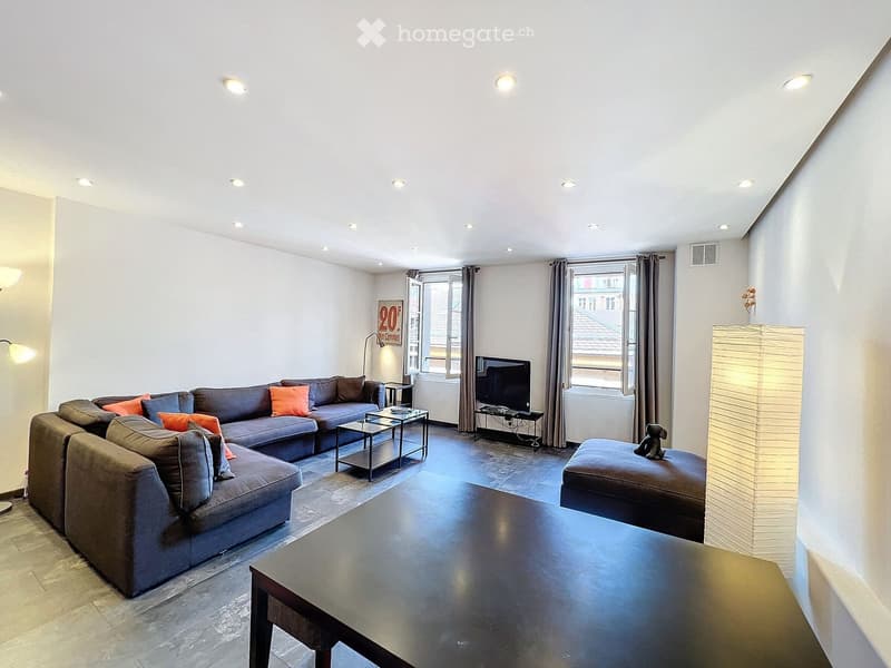 Modern living room with grey sectional sofa, orange throw pillows, coffee table, ottoman, and a television on a stand. The room features large windows with curtains and recessed ceiling lighting.