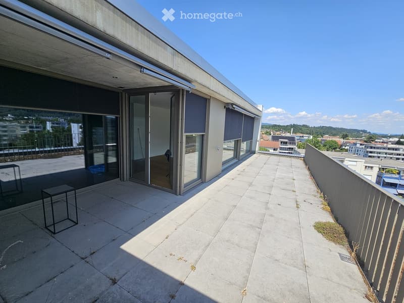 Modern rooftop terrace with concrete flooring, glass sliding doors leading to an indoor space, and a cityscape view.