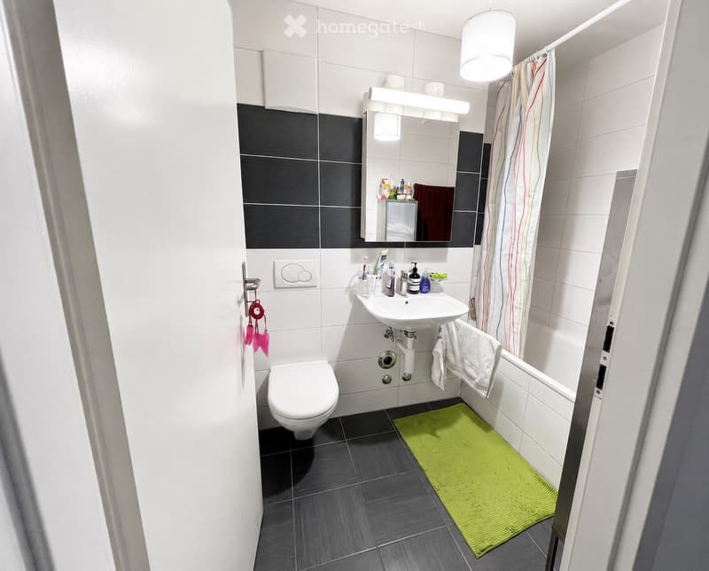 Modern bathroom with white toilet, sink, mirror, and a green bath mat. Features dark grey tiles and a white shower curtain.