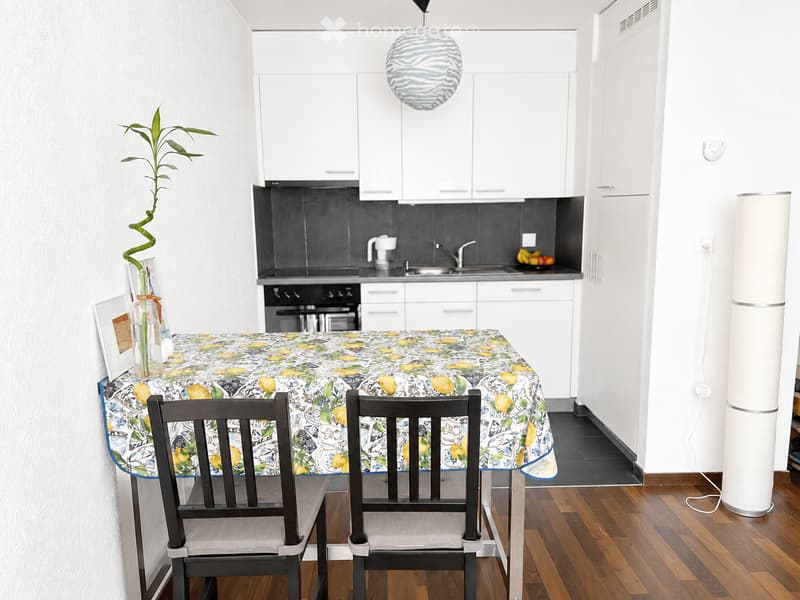 Modern kitchen with white cabinets, a round dining table with lemon-patterned tablecloth, two chairs, and a hanging lamp. The kitchen features a black backsplash, stainless steel appliances, and a small potted plant on the table.