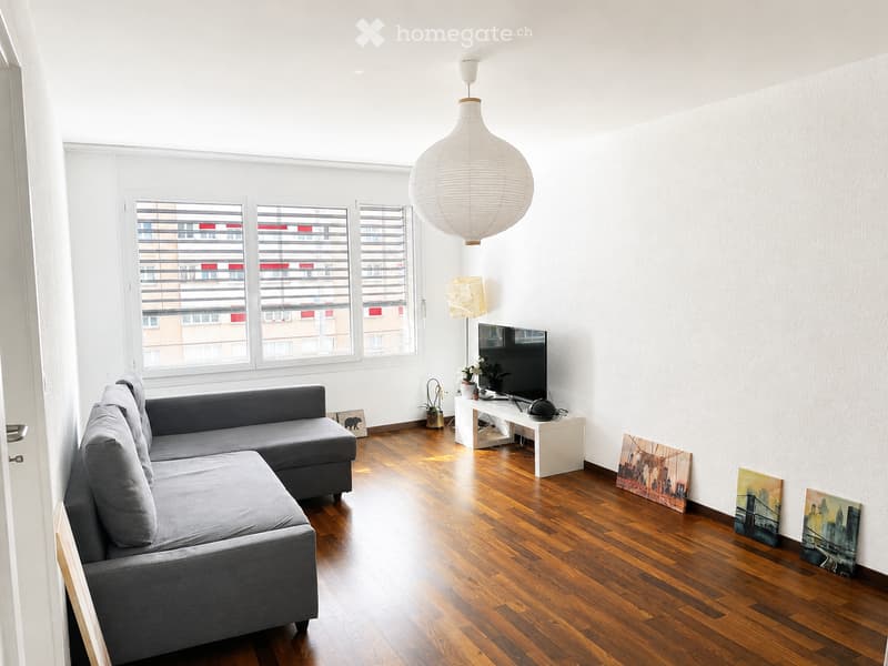 Modern living room with wooden flooring, large windows, grey sectional sofa, coffee table, TV, and decorative artwork.