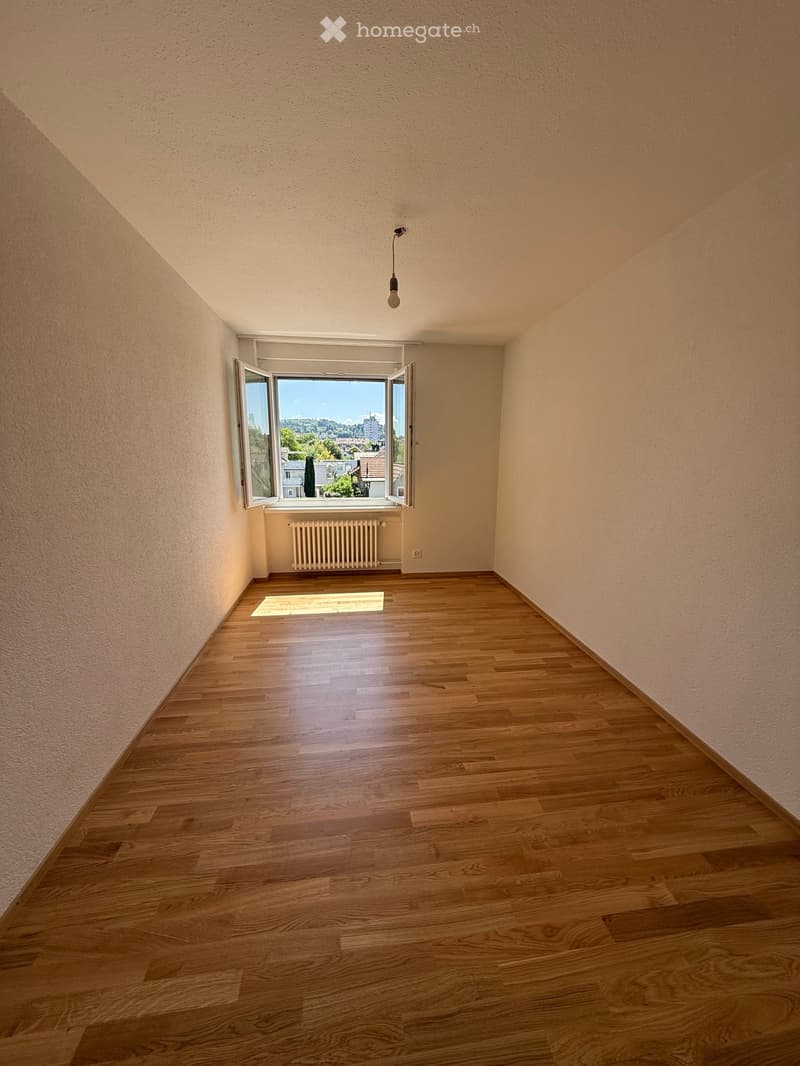 Empty room with wooden flooring, light-colored walls, and a large window with open shutters, allowing natural light to enter. A radiator is visible below the window, and a single pendant light hangs from the ceiling.