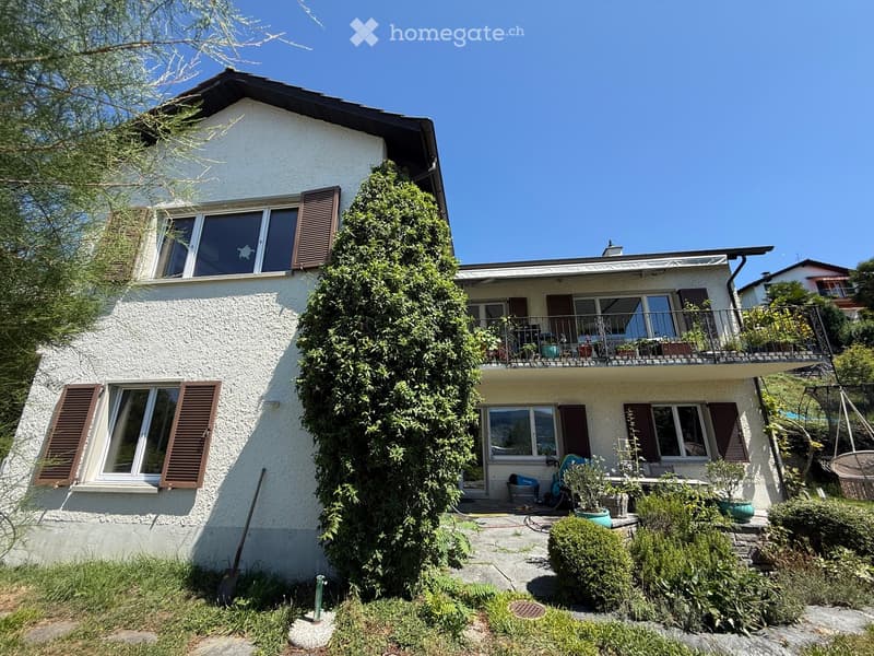 Two-story house with white exterior, multiple windows with brown shutters, balcony on the upper level, and surrounding greenery.
