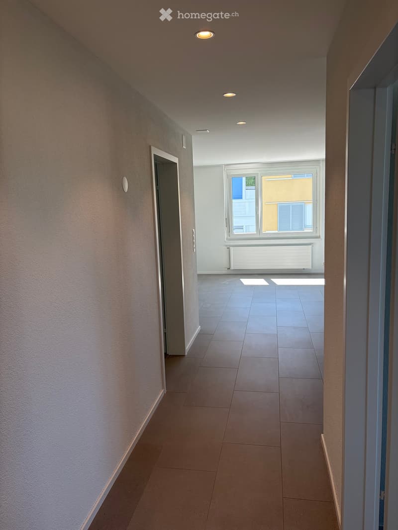 Empty hallway with tiled floor, white walls, recessed lighting, and a window at the end showing a view of a building.