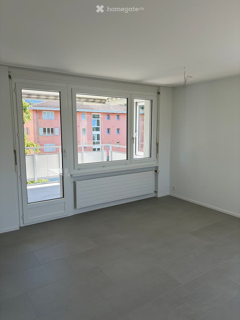 Empty room with grey tiled floor, white walls, large window with white frame, radiator, and a sliding door leading to a balcony.